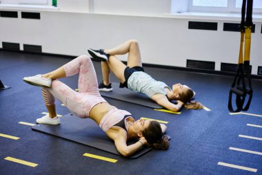 Vücut yüksekliği, spor kıyafetleri giymiş kadın sporcuların elleri arkada, paspaslar üzerinde köprü egzersizi yapmaları.