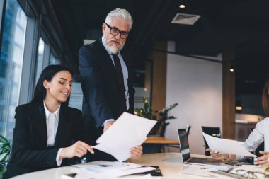 Bir grup resmi kıyafetli iş arkadaşı dizüstü bilgisayar kullanıyor ve iş kuruyor. Bu arada erkek patron da modern ofisteki kağıtlara bakıyor.