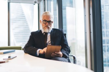 Resmî takım elbise ve gözlük takan olgun sakallı adam tableti incelerken kameraya bakıyor ve not defteriyle masanın yanında tek başına oturuyor.