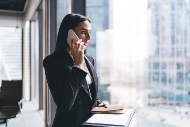 Konsantre kadın girişimci resmi kıyafetler içinde cam duvarın yanında cep telefonuyla konuşurken ve şehir merkezine bakarken not defteriyle duruyor.