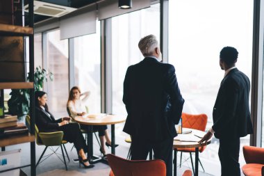 Ofis kafeteryasındaki ahşap masaların yanında duran ve iş projelerini tartışan resmi giyinmiş bir grup erkek ve kadın meslektaşız.