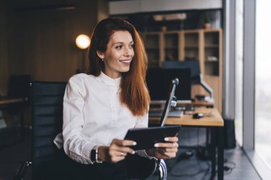 Olumlu genç bayan resmi giysileri gülümseyerek ve başka tarafa bakarak modern çalışma alanlarında çalışma saatlerinde tablet kullanarak otururlar.