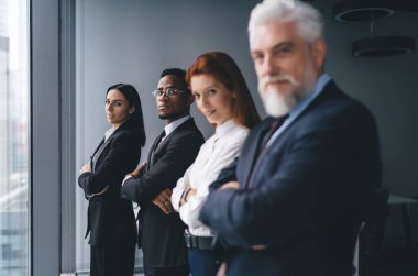 Modern ofis odasındaki cam duvarın yanında duran ve ofiste vakit geçirirken kameraya bakan çok ırklı iş adamları.