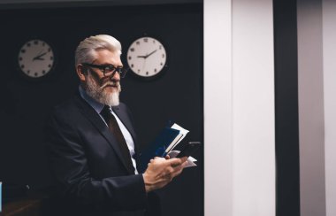 Pozitif sakallı, iyi giyimli, gözlüklü, evrak işleriyle uğraşan, duvar kenarında saatli ve cep telefonu ekranına bakan bir patron.