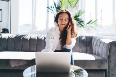 Evde boş zamanlarında akıllı telefondan arkadaşınla konuşurken bilgisayarın ekranına bakan konsantre genç İspanyol kadın iş kadını.