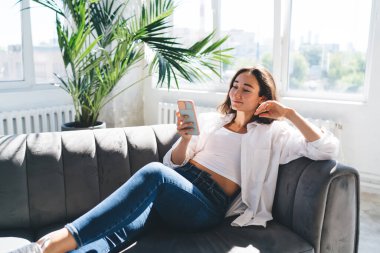 Oturma odasında boş zamanlarında kanepede dinlenip mesajlar okurken gülümseyen ve akıllı telefon ekranına bakan genç etnik kadın.