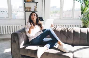 Çağdaş apartmandaki jest gibi kameraya bakarken kanepeye oturmuş dizüstü bilgisayarını karıştıran etnik kökenli bir kadın.