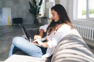 Uzun siyah saçlı, pozitif etnik bir kadının geniş bir odada kanepede otururken günlük kıyafetlerini giyerek netbook 'a mesaj yazmasının yan görüntüsü.