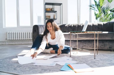 Uzun siyah saçlı, rahat bir Asyalı kadın. Günlük giysiler giyiyor. Evde çalışırken yerde kağıtlarla oturuyor.