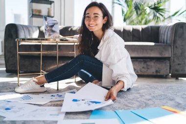 Sırıtkan Asyalı kadın serbest çalışan, uzun siyah saçlı, günlük kıyafetli, elinde belgelerle yerde oturan ve kameraya bakan.