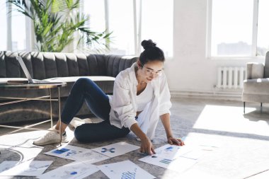 Bütün vücudu gözlüklü genç bir kadın dağınık belgelerle yerde oturuyor ve evdeki oturma odasında uzaktan kumandalı bir iş projesi üzerinde çalışıyor.