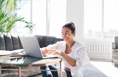 Gülümseyen Asyalı kadın serbest kıyafet ve gözlük takıyor küçük masada oturuyor ve oturma odasında dizüstü bilgisayarına bilgi yazıyor.
