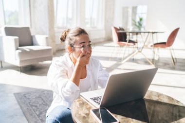 Gözlüklü genç bir kadın masada oturmuş, elleri ağızlarını kapatırken evdeki oturma odasında uzaktan kumandalı bir iş projesi için bilgisayar kullanıyor.