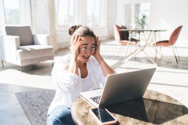 Evde oturma odasında netbook kullanarak uzak bir proje üzerinde çalışırken gündelik kıyafetleriyle masada oturup kafaya dokunan mutlu kadın serbest yazar.