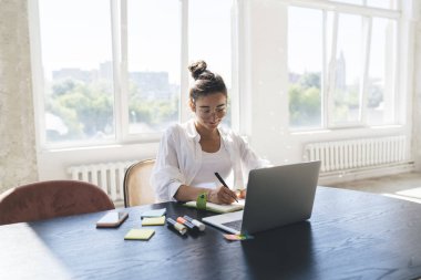 Gözlüklü mutlu genç bir kadın evde oturma odasında dizüstü bilgisayar kullanarak üniversite ödevini yaparken kırtasiye malzemesi ve planlamasıyla masada oturuyor.