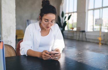 Günlük kıyafetli ve gözlüklü bir kadın ahşap masada oturuyor ve evde dinlenip ekrana bakarken cep telefonunu karıştırıyor.