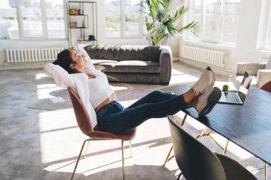 Genç bir bayanın rahat koltukta rahat rahat oturması, ellerinin başının arkasında olması ve bilgisayarın masanın üzerinde kırılıp gerilmesi.