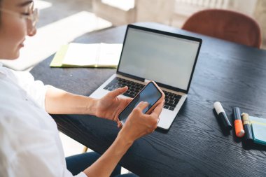 Yüksek açılı kadın serbest yazar, ahşap masada netbook ile oturuyor ve cep telefonunu ekranda tarıyor.