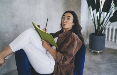 Etnik kadın ofis çalışanlarının evde boş zamanlarında bacak bacak bacak üstüne atıp kameraya bakarken tatmin olmalarının yan görüntüsü.