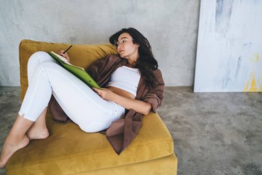 Genç, etnik bir bayanın rahat bir kanepede yatıp boş zamanlarında evde dinlenirken kalemle not defterine not almasından ve çalışma programı planlamasından.