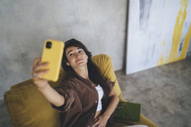 Uzun saçlı, rahat ve gülümserken cep telefonundan selfie çeken genç etnik bir kadın.