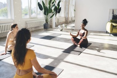 Mat üzerinde oturan ve sabahları hafif stüdyoda yoga sırasında Padmasana pozu veren çok ırklı bayan arkadaşların yan görüntüsü.