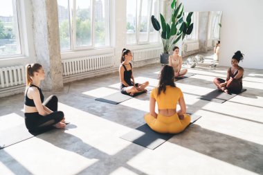 Etkinlik giysileri içinde bir grup kadın arkadaş minderlerin üzerinde oturuyor ve yoğun güneş ışığı altındaki stüdyoda yoga dersinde birlikte poz veriyorlar.