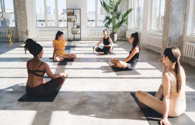 Yoga antrenmanında yerde dinlenirken ve birlikte boş vakit geçirirken birbirine bakan ve konuşan etnik çeşitliliğe sahip bir grup kadın.