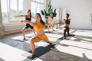 Sportif giysiler içinde, savaşçı pozunda yoga yapan ve minderlerin üzerinde duran neşeli genç bir kadın vücudu.