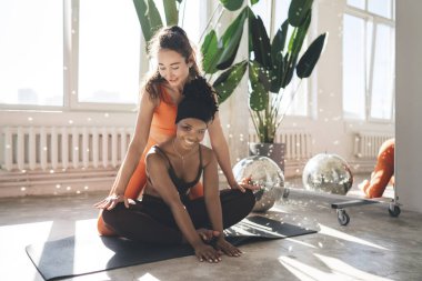 Gülümseyen Afro-Amerikan kadın tüm vücudu yoga pozisyonunda elleri yerde oturmuş kameraya bakarken İspanyol kadın antrenör antrenman sırasında bacaklarını yerleştiriyor.