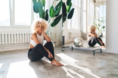 Spor kıyafetleri giyen sakin bir bayan sporcunun, yoga stüdyosunda antrenman yaptıktan sonra gülümsemesi ve büyük aynanın yanında oturması.