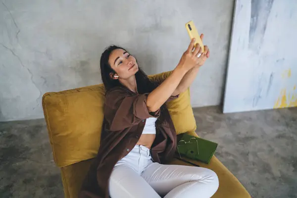 stock image High angle of young female with earbuds in casual wear sitting and using mobile phone while resting on comfortable armchair