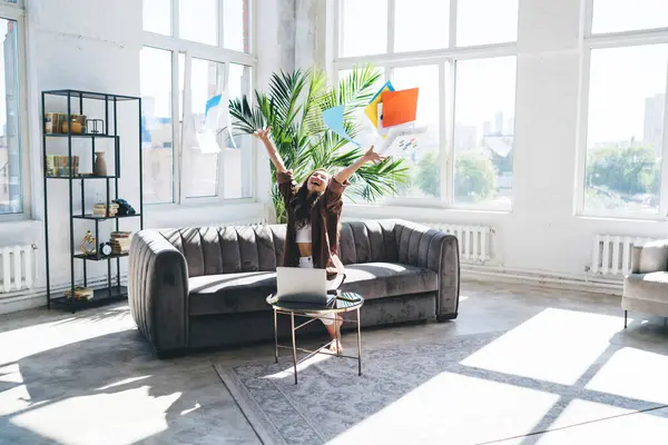 stock image Rejoicing female entrepreneur throwing work papers up to show victory in project goals accomplishment while sitting on sofa in spacious living room