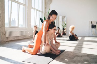 Genç, gülümseyen bir bayanın lotus pozisyonunda yoga yapması ve etnik kız arkadaşının spor salonunda boş vakit geçirmesi ve ellerini dizlerinin üstüne koyup oynaması.