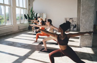 Warrior pozunda duran genç kadın grubunun yan görüntüsü ya da büyük pencereli yoga seansı sırasında minderlerin üzerinde duran Virabhadrasana