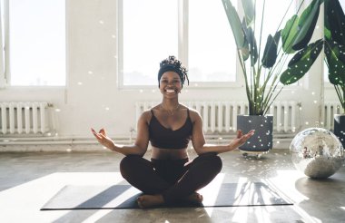 Spor kıyafetli, Lotus pozunda gülümseyen, kameraya bakan ve yoga yaparken meditasyon yapan zayıf Afrikalı Amerikalı bir kadın.