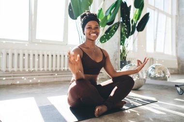 Spor kıyafetli, lotus poz vermiş, elleri Gyan Mudra 'da oturmuş gülümseyip kameraya bakarken meditasyon yapan genç siyahi bir kadın.