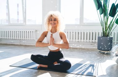 Anjali Mudra 'da mindere oturmuş gülümseyen genç bir kadın vücudu ya da selam veren fok pozisyonu ve kameraya bakarken yoga çalışması.