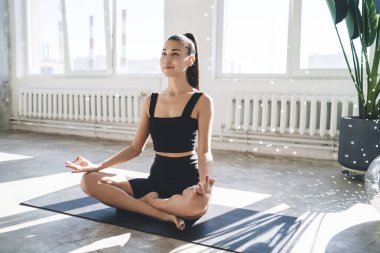 Lotus pozisyonunda otururken ya da Padmasana 'da güneşli bir günde modern stüdyoda yoga yaparken yüzünü çeviren genç bir kadın.