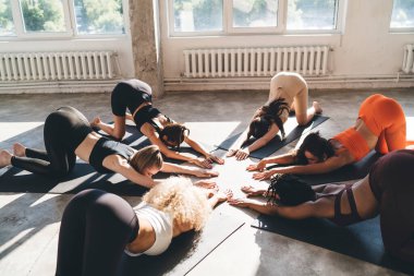 Aktif giyimli bir grup genç kadın paspaslar üzerinde uzanıp modern stüdyoda yoga yaparken Uttana Shishosana pozu veriyorlar.