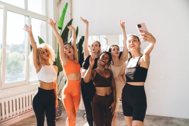 Spor kıyafetleri içinde, pencerenin yanında elleri havada dururken akıllı telefondan selfie çeken bir grup mutlu çok ırklı kadın.