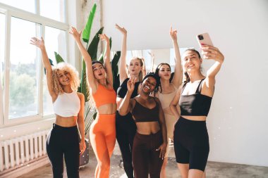 Antrenmanlı, dar elbiseli, spor salonunda gülümseyen ve el kaldıran, cep telefonuyla selfie çeken mutlu atletik kadınlar.