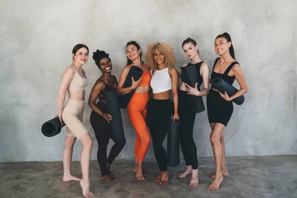 stock image Full length of young multiracial women in sportswear standing together with fitness mats and looking at camera during fitness workout in gym