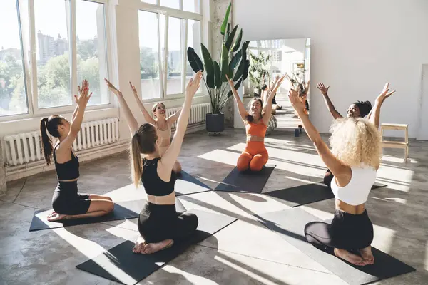 stock image Full body of group of barefoot young sportswomen in activewear sitting on mats in circle and taking Vajrasana pose with arms raised while practicing yoga together during class in studio