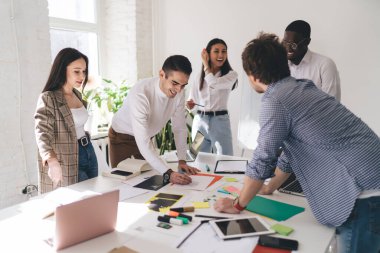 Gündelik giysiler içinde bir grup çok ırklı iş arkadaşı, modern ofiste başlangıç fikirleri üzerinde çalışırken evrak ve belgelerle birlikte masada toplanıyor.
