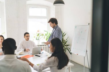 Genç adam elinde kağıtlarla elinde çok ırklı meslektaşlarıyla toplantı masasında dizüstü bilgisayar kağıtları ve kalemlerle proje detaylarını tartışırken...
