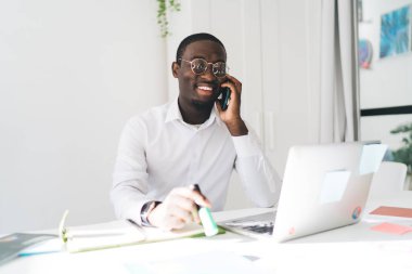 Pozitif Afro-Amerikan erkek girişimci elinde laptop kağıtları ve yapışkan notlarla masada otururken elinde kalemle cep telefonuyla güneşli ofis odasında konuşuyor.