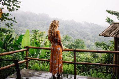Yeşil bitki ve ağaçlarla dolu tropikal bahçedeki misafir evinin ıslak ahşap tahtasında bambu çitlerin yanında duran tanınmayan kadın turistin arka manzarası.