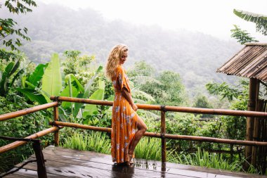 Tahta terasta duran ve yeşil egzotik ağaçların yanında dinlenirken aşağıya bakan zarif, genç bir kadın vücudu.