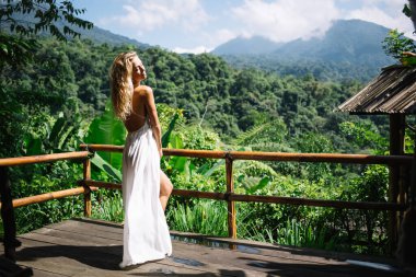 Beyaz elbiseli, uzun sarı saçlı, rahat genç bir kadın, ahşap terasta çitin yanında dikilirken yeşil dağların manzarasının keyfini çıkarıyor.
